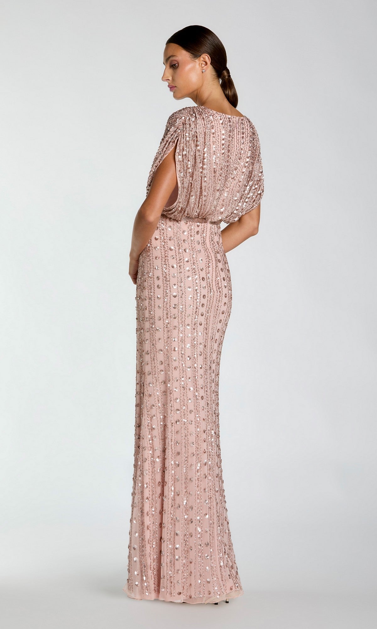 A woman stands in profile against a plain light background wearing the Long Formal Dress: Mac Duggal 23081—a floor-length, rose gold sequin beaded gown featuring short sleeves and a fitted silhouette.